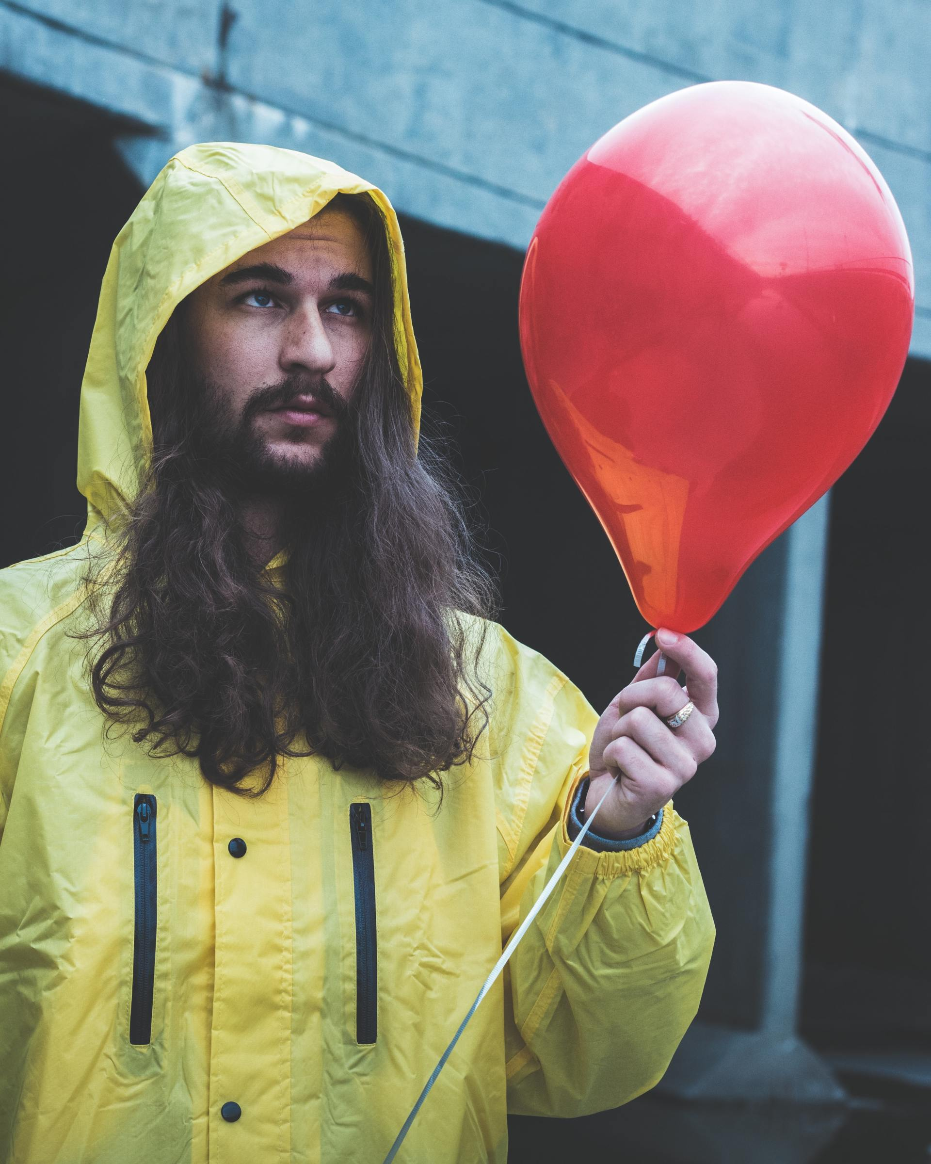 A man with long dark hair and a moustache wears a yellow rain jacket and holds a red balloon