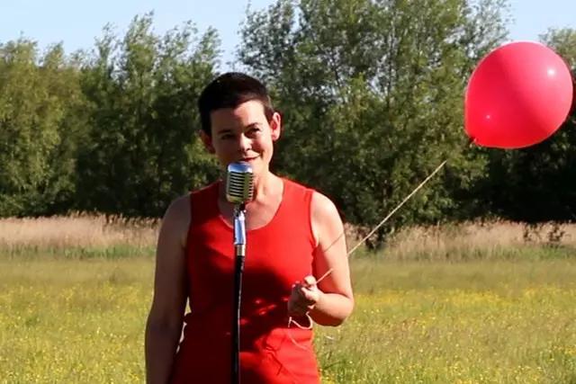 Anna Tolputt speaks into a condenser microphone in a field. She wears a red dress and holds a red balloon.